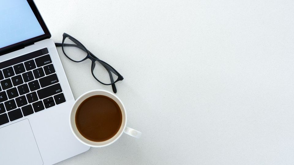 Laptop, Brille und Kaffeetasse auf einem hellen Schreibtisch von oben